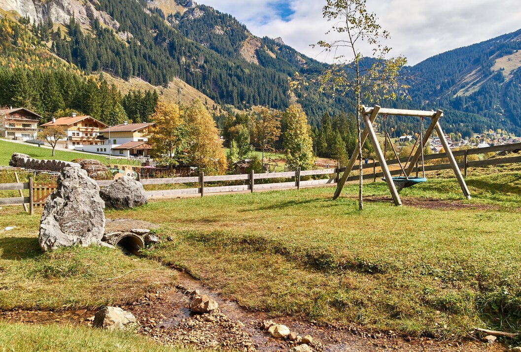Spielplatz mit Schaukel, Bachlauf, Holzzaun und Häusern vor bewaldeten Bergen