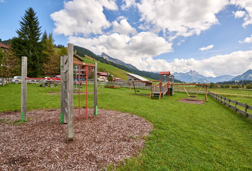 Spielplatz mit Klettergerüst, Schaukeln und Rutsche vor Bergkulisse unter blauem Himmel mit Wolken