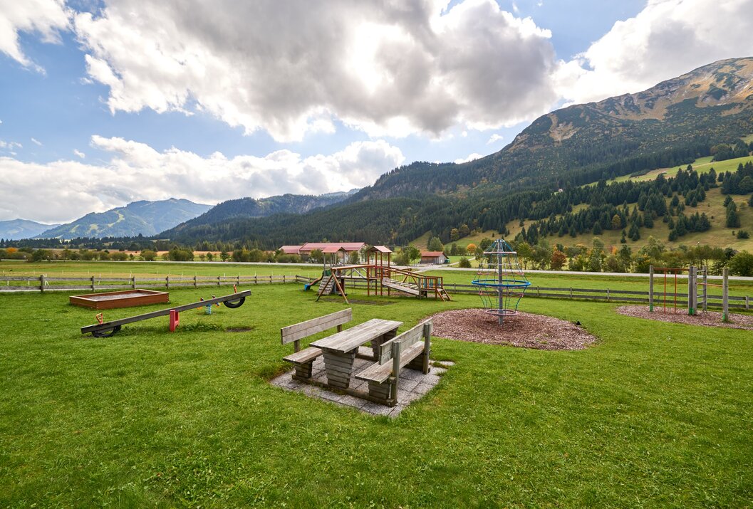 Spielplatz mit Tisch, Bänken, Schaukel, Wippe und Klettergerüst vor Bergkulisse