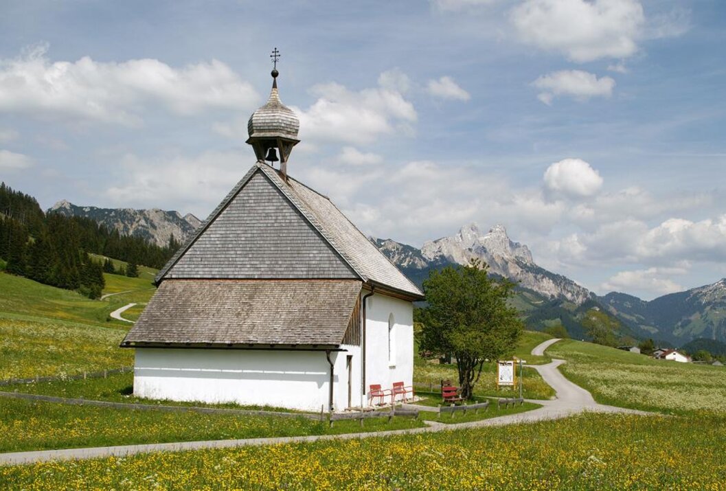 Kleine weiße Kapelle auf grüner Wiese vor Bergkulisse unter blauem Himmel