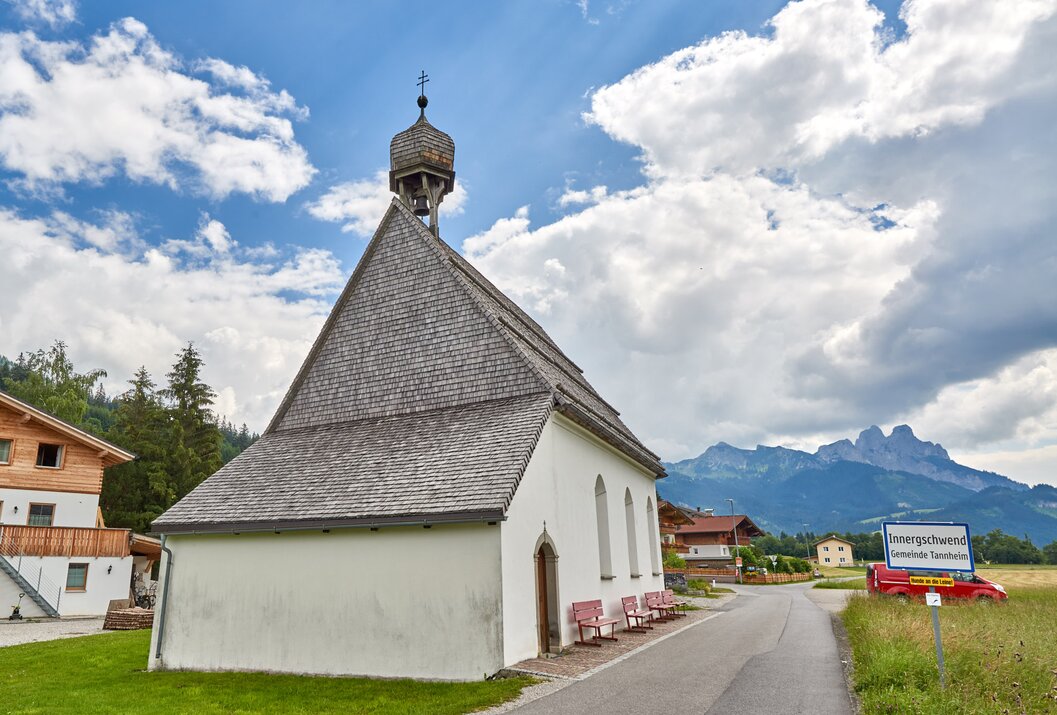 Weiße Kapelle mit grauem Dach vor blauem Himmel mit Wolken und Bergen im Hintergrund