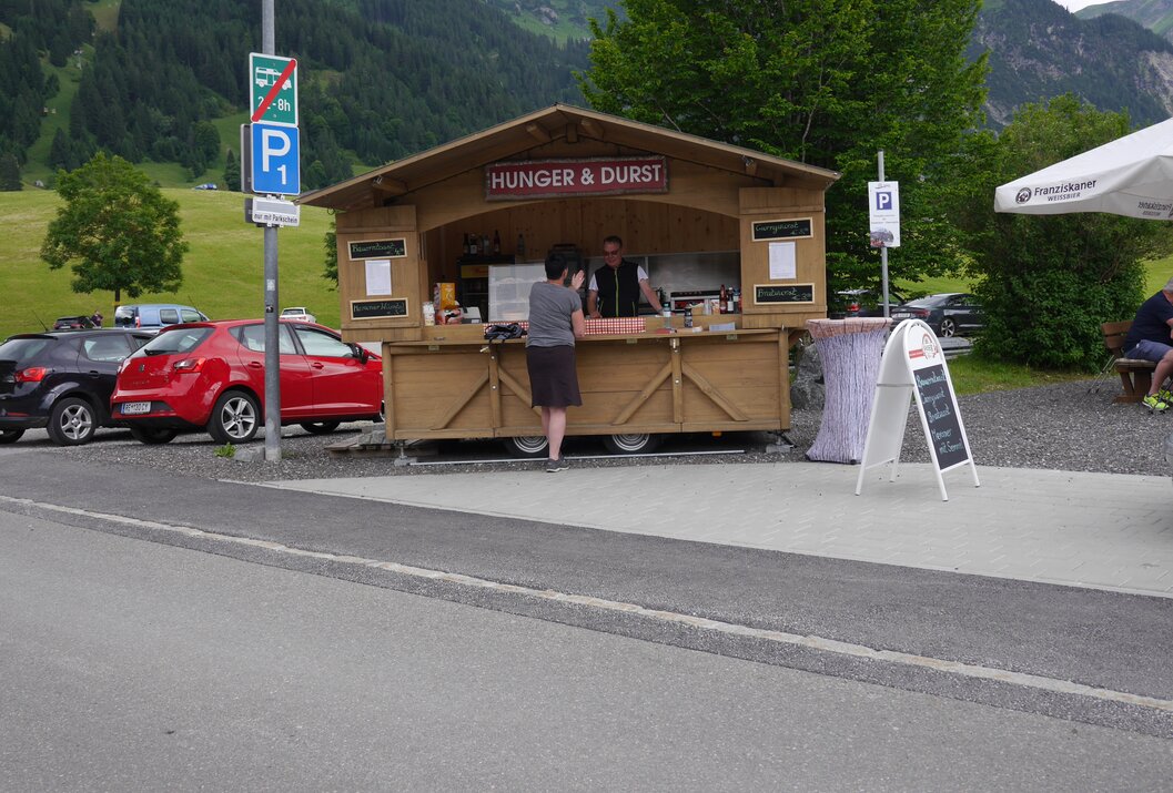 Imbissstand "Hunger & Durst" mit Kundin vor grüner Wiese und Bergen