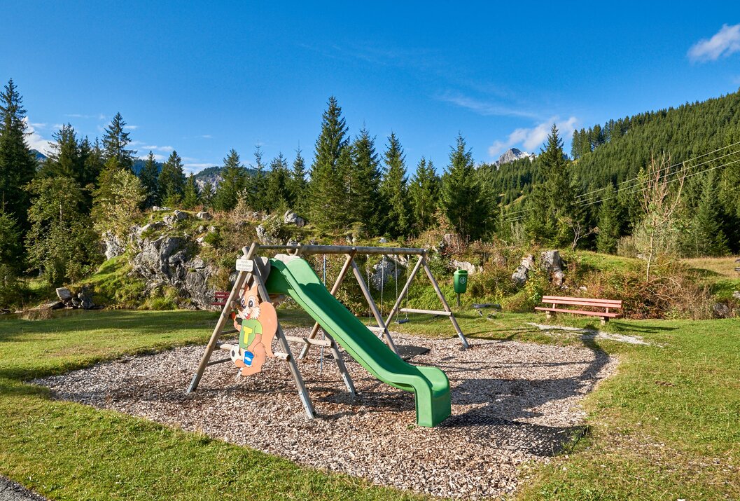 Spielplatz mit Rutsche, Schaukeln und Holzboden vor bewaldetem Hügel unter blauem Himmel