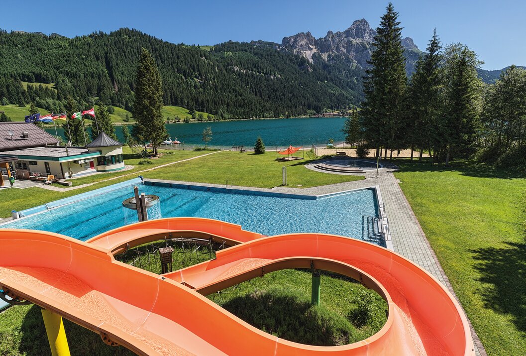 Swimming pool with slide in front of mountain lake and wooded mountains