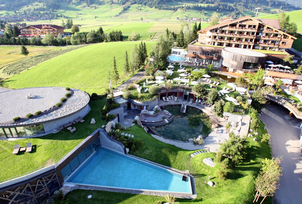 Luftaufnahme eines Resorts mit Pools, Liegen und Grünflächen vor bergiger Landschaft
