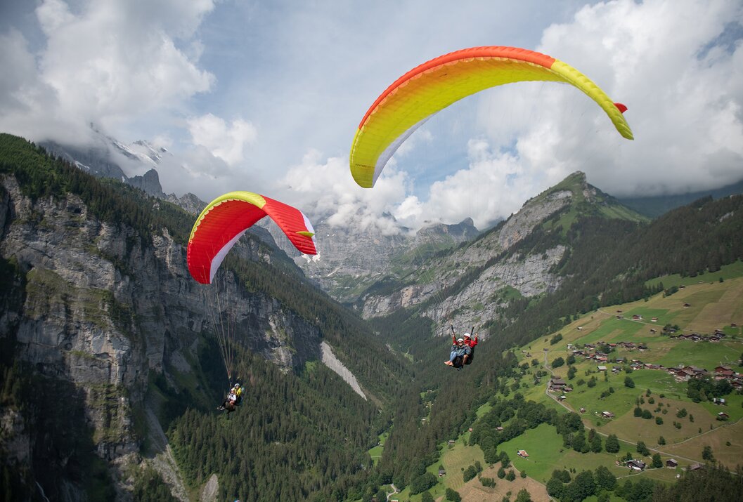 Zwei Gleitschirmflieger vor Berglandschaft mit grünen Hängen und bewölktem Himmel