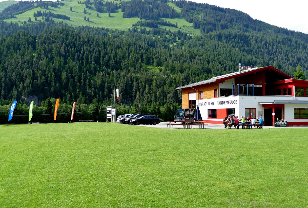 Gebäude mit Aufschrift "Paragliding Tandemflüge" vor grüner Wiese und bewaldetem Berg
