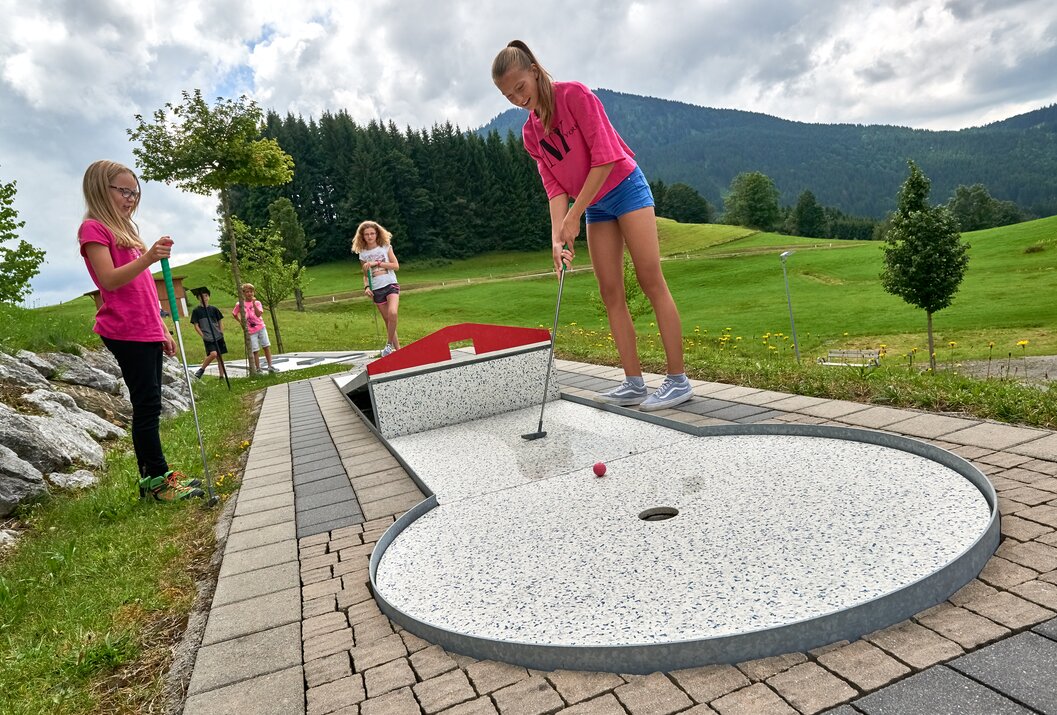 Mädchen spielt Minigolf, andere Kinder im Hintergrund, Berglandschaft