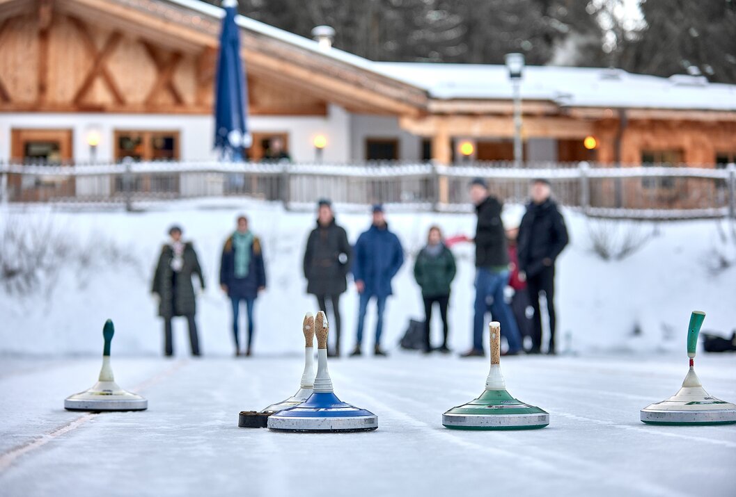 Curlingsteine auf Eisfläche, im Hintergrund verschwommene Menschengruppe vor einem Gebäude