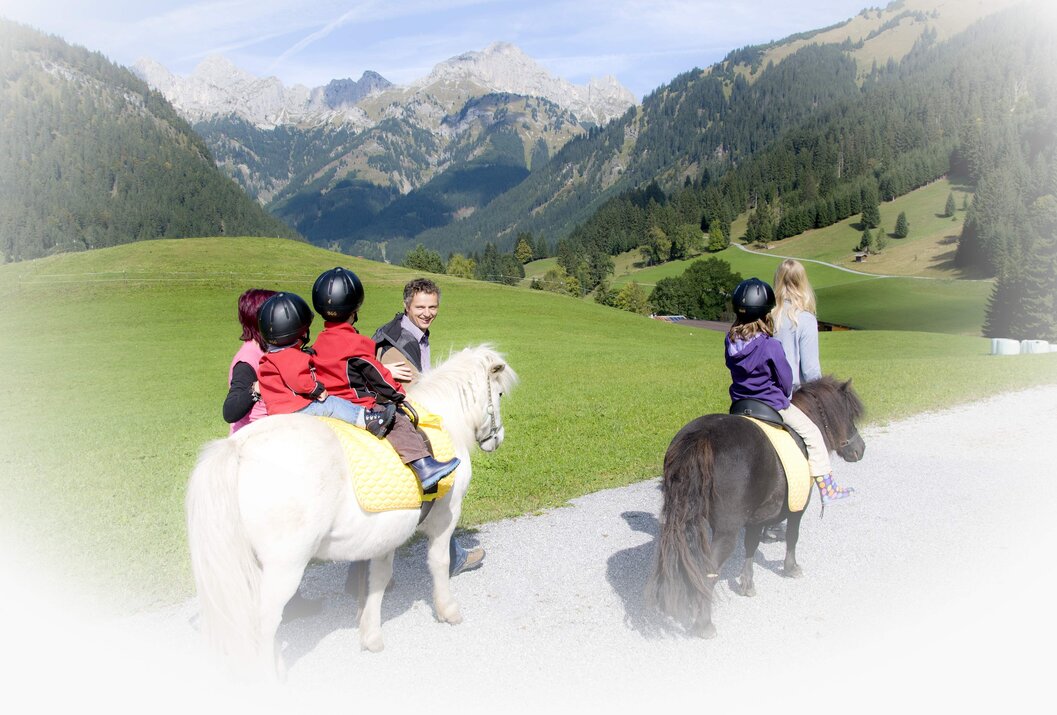 Kinder reiten auf Ponys vor Bergkulisse mit Betreuer