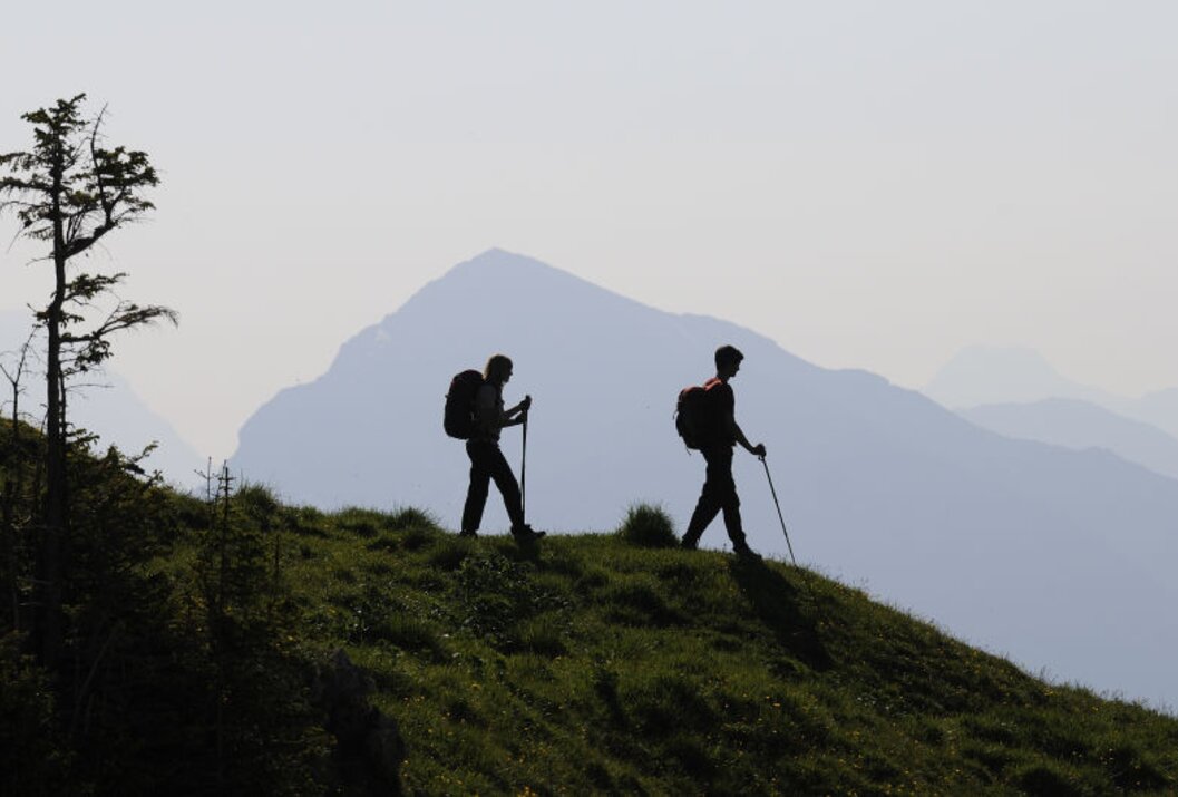 Zwei Wanderer mit Rucksäcken und Stöcken auf einem Hügel vor Bergkulisse als Silhouetten