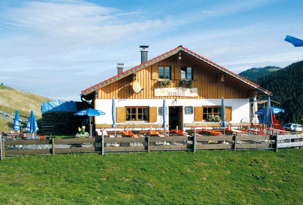 Holzhaus mit braunen Fensterläden und Außengastronomie auf einer grünen Wiese