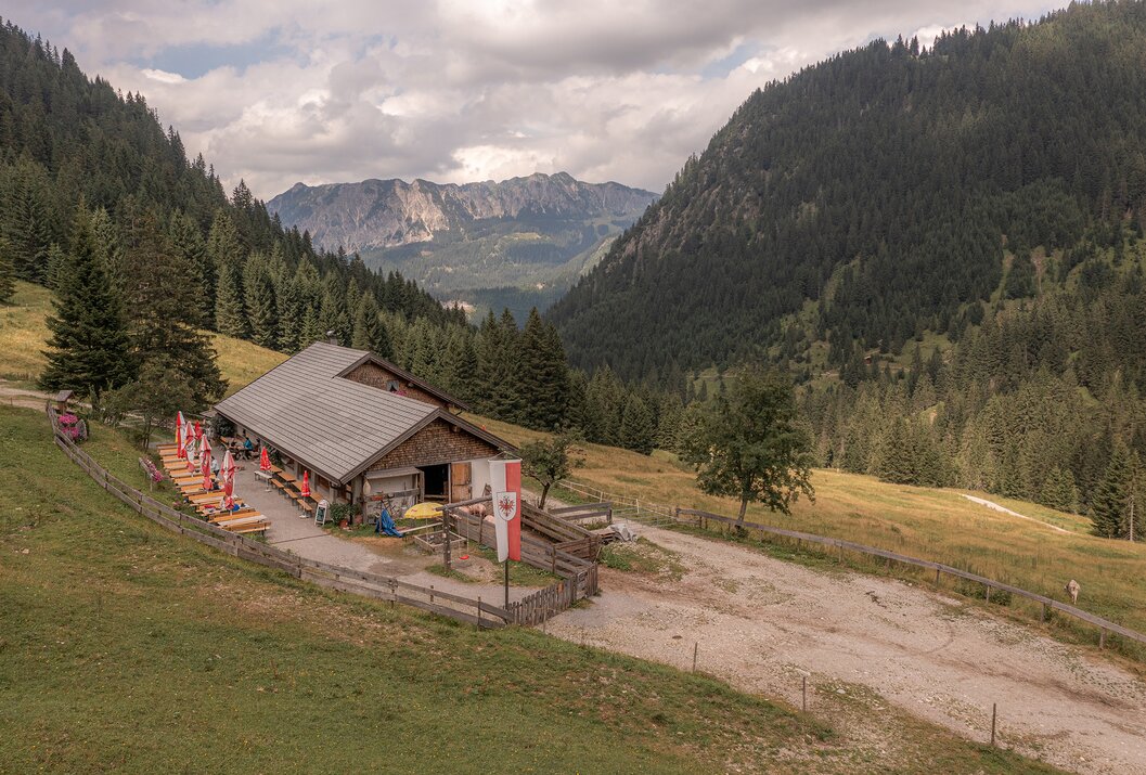 Hütte in Berglandschaft mit Wald, Wiesen und Bergen im Hintergrund unter bewölktem Himmel