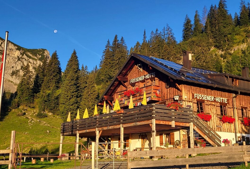 Hölzerne Berghütte "Füssener Hütte" mit gelben Sonnenschirmen vor bewaldetem Hang unter blauem Himmel