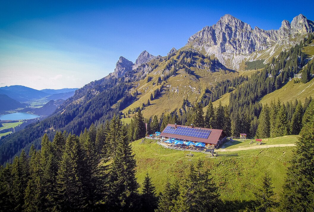 Berghütte mit blauen Sonnenschirmen vor bewaldetem Hang und Bergmassiv unter blauem Himmel
