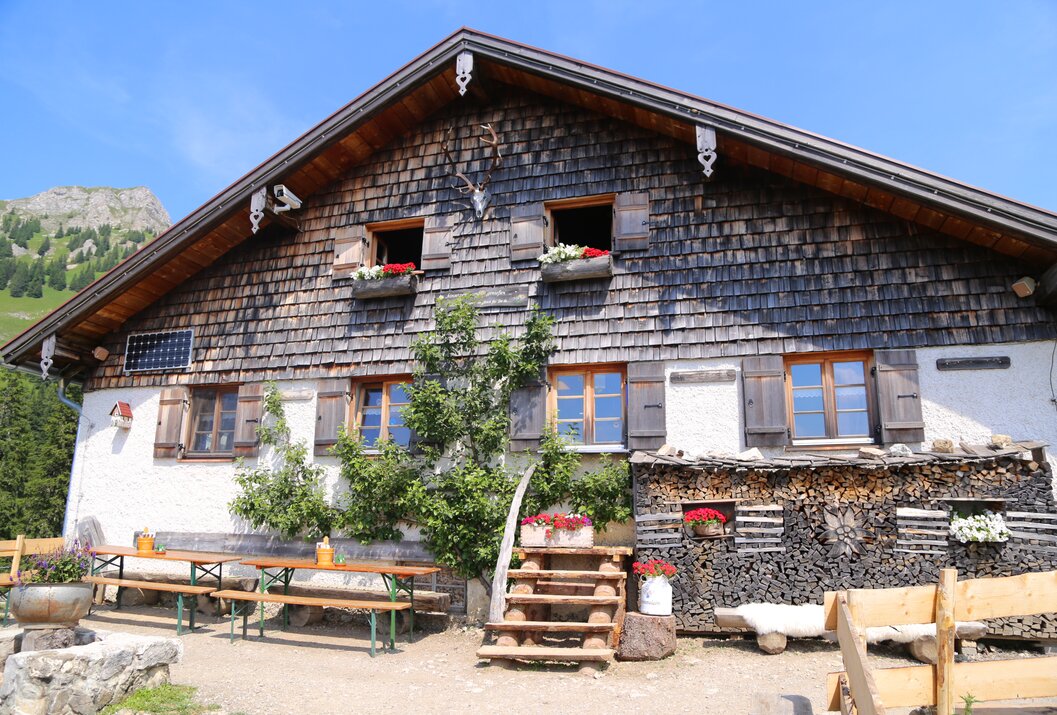 Altes Holzhaus mit Schindelfassade, Fenster mit Blumenkästen, Holzstapel und Sitzgelegenheiten vor dem Haus