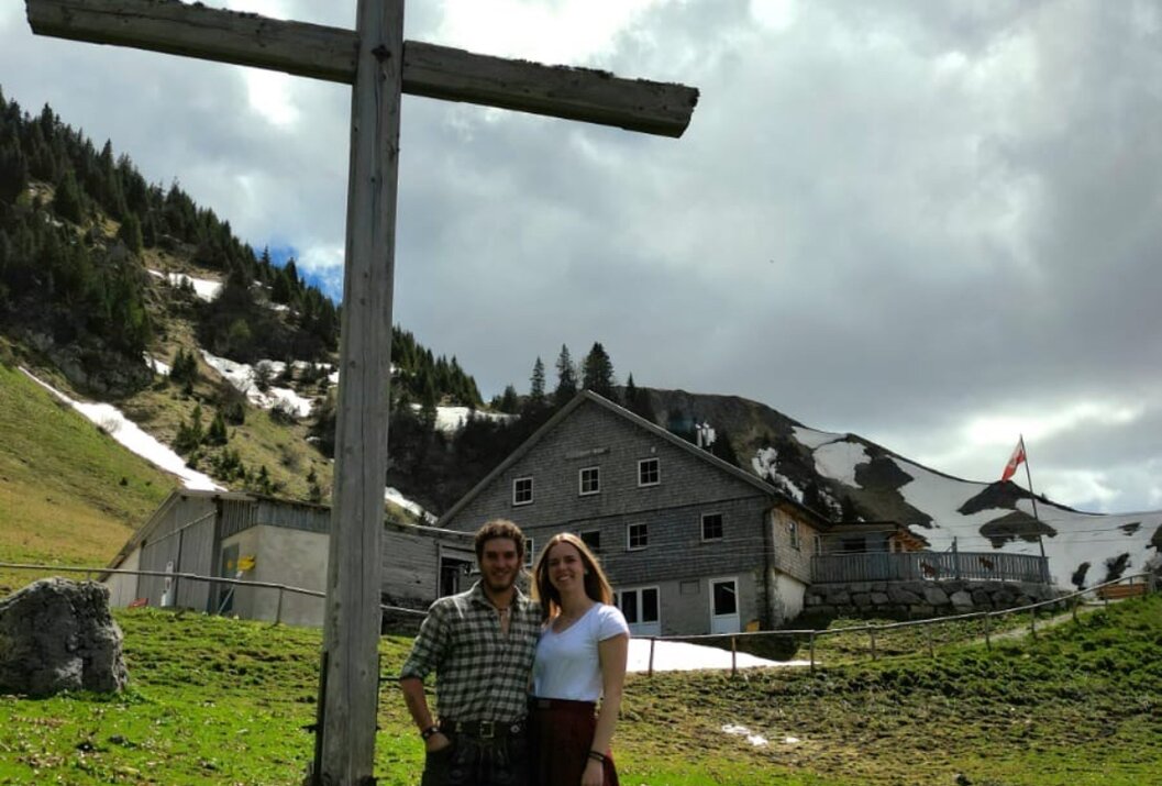 Paar in Tracht vor Holzkreuz, Hütte und Berglandschaft mit Schnee im Hintergrund