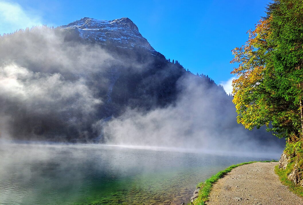 Bergsee mit Nebel, Berg im Hintergrund, Baum rechts, blauer Himmel