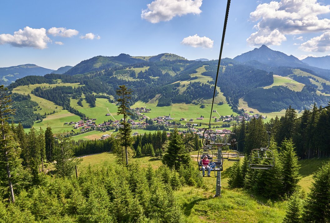 Sessellift mit zwei Personen vor Berglandschaft mit grünen Wiesen und bewaldeten Hängen