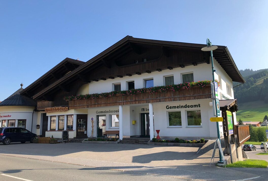 Gemeindeamt mit dunklem Dach und Blumenkästen unter blauem Himmel