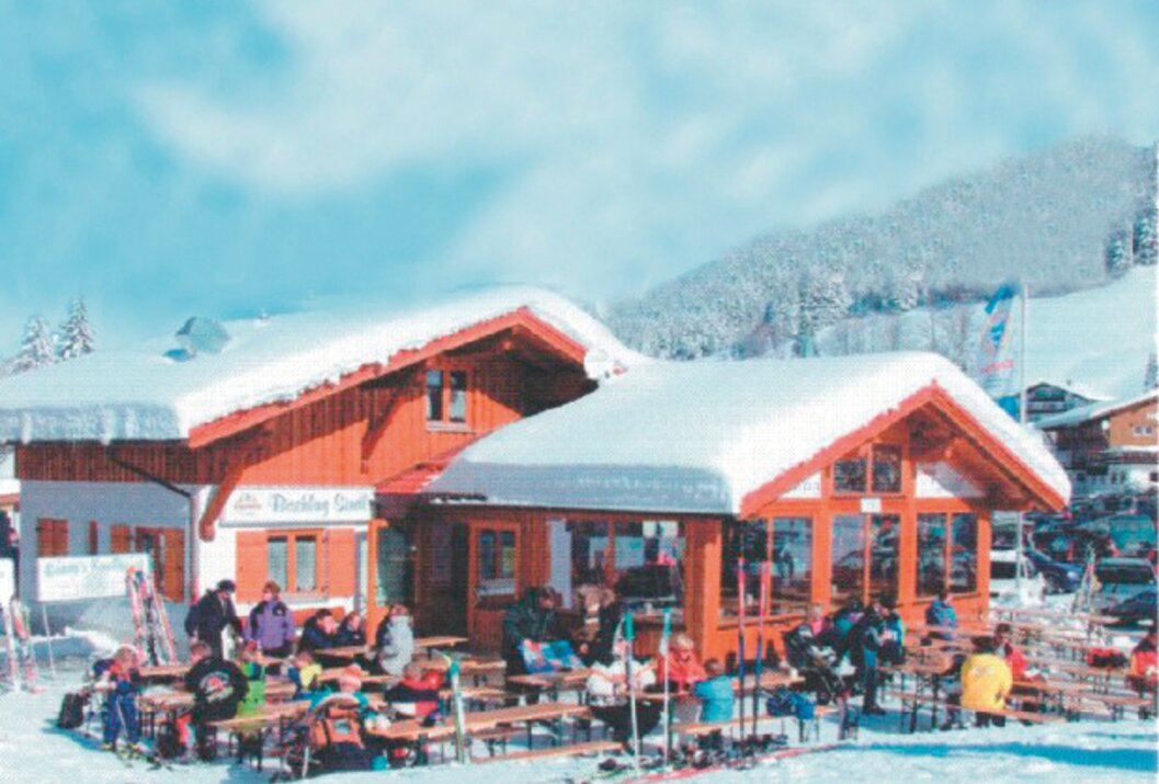 Verschneite Berghütte mit Gästen auf der Terrasse vor schneebedeckter Berglandschaft