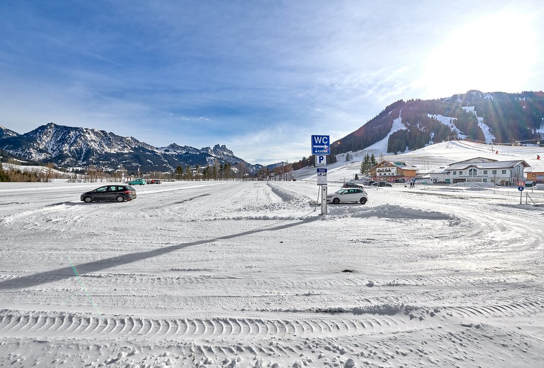 Verschneiter Parkplatz mit WC-Schild vor Bergkulisse unter blauem Himmel