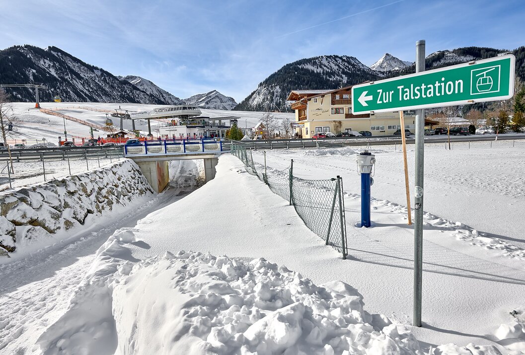 Winterlandschaft mit Schnee, Brücke, Schild "Zur Talstation" und Bergen im Hintergrund