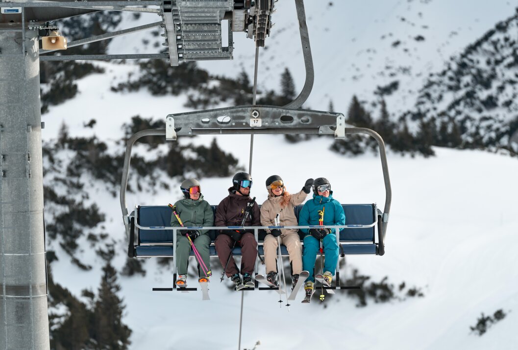 Vier Skifahrer sitzen in einem Sessellift vor verschneiter Landschaft