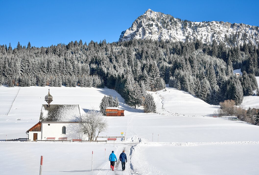 Winterlandschaft mit schneebedeckter Kapelle, Wald und Berg im Hintergrund, zwei Personen im Vordergrund