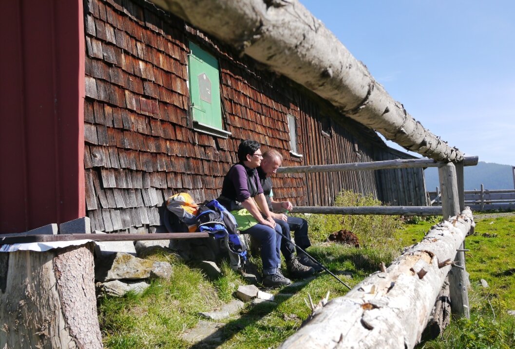 Two hikers rest in front of a wooden hut in the sunshine