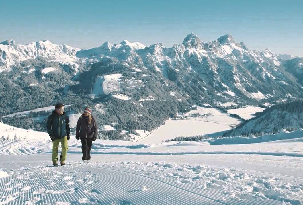Zwei Personen auf Skipiste vor verschneiter Bergkulisse